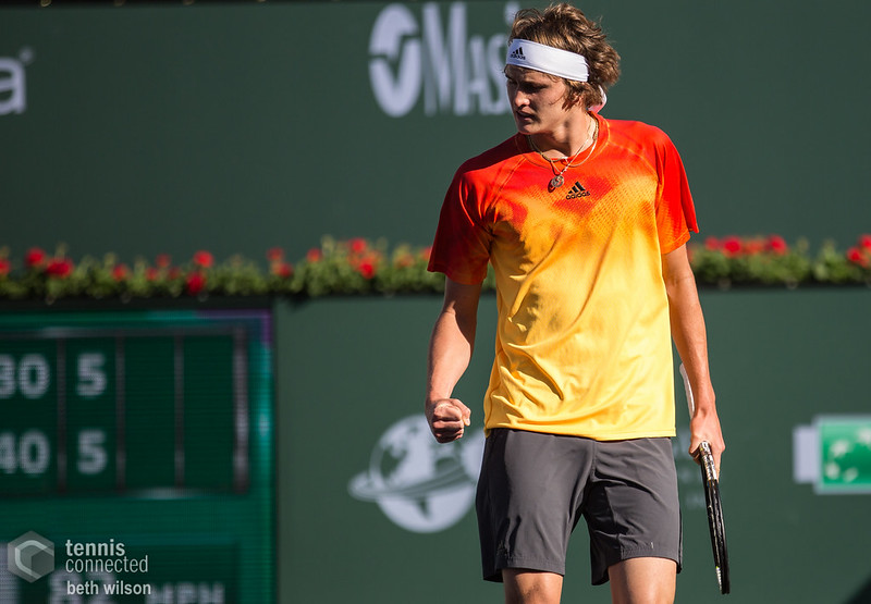 Zverev con camiseta naranja y amarilla, pantalón gris, raqueta en la mano izquierda y cinta blanca en el pelo